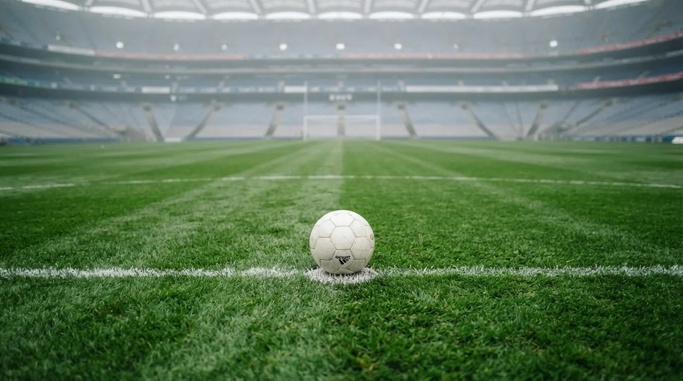Ein Fußball liegt auf dem Elfmeterpunkt eines leeren Rasenplatzes, im Hintergrund eine Tribüne im Dunst.