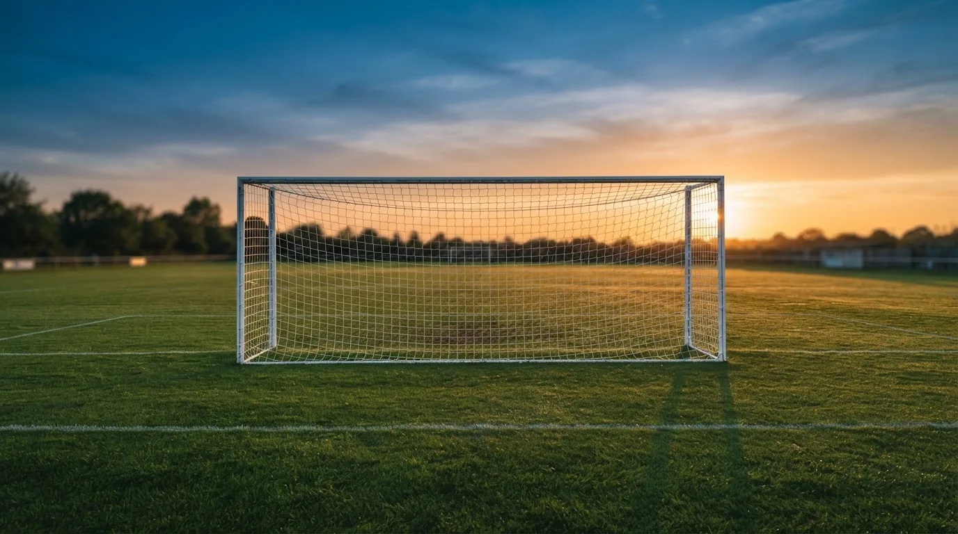 Ein Fußballtor mit Netz bei Sonnenuntergang auf einem leeren Platz, die Schatten der Torpfosten liegen lang auf dem Gras.
