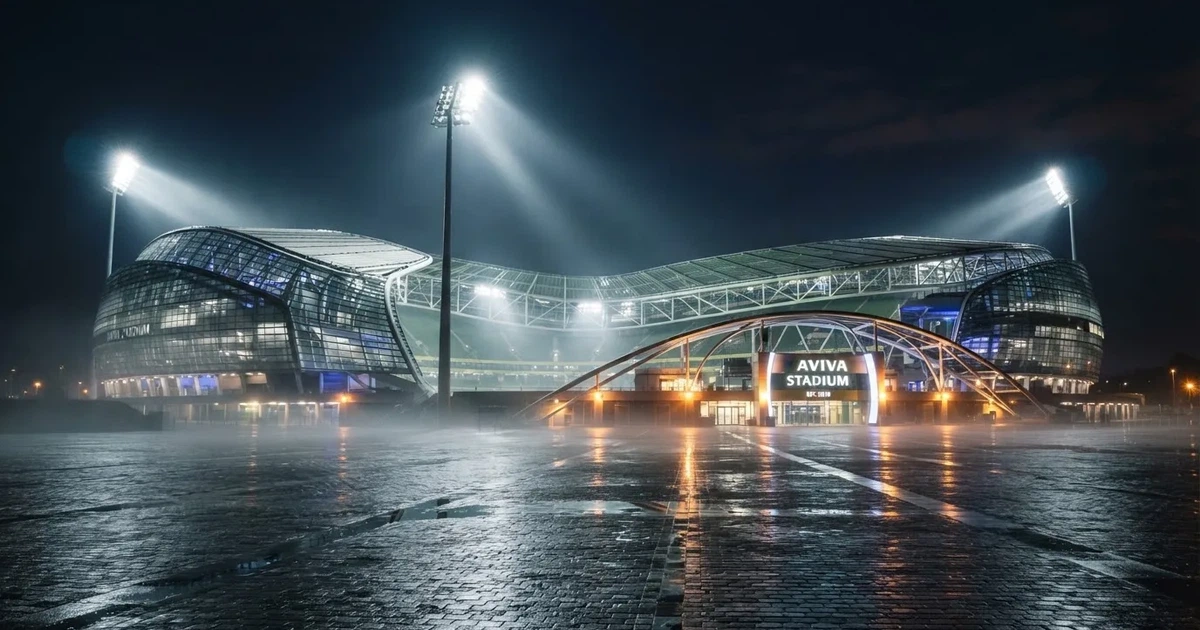 Ein großes Fußballstadion bei Flutlicht von außen, davor ein leerer Vorplatz mit nassem Pflaster und Dunst.
