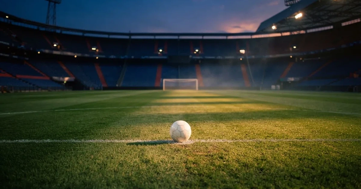 Ein Fußball liegt auf dem Anstoßpunkt eines leeren Rasenplatzes, im Hintergrund eine große Stadiontribüne im Abendlicht.