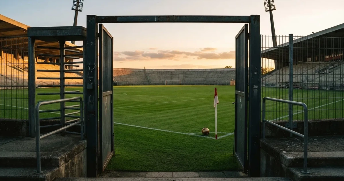 Blick durch ein Stadiontor auf einen grünen Rasen im Abendlicht, ein Ball ruht an der Eckfahne, die Tribünen sind leer.