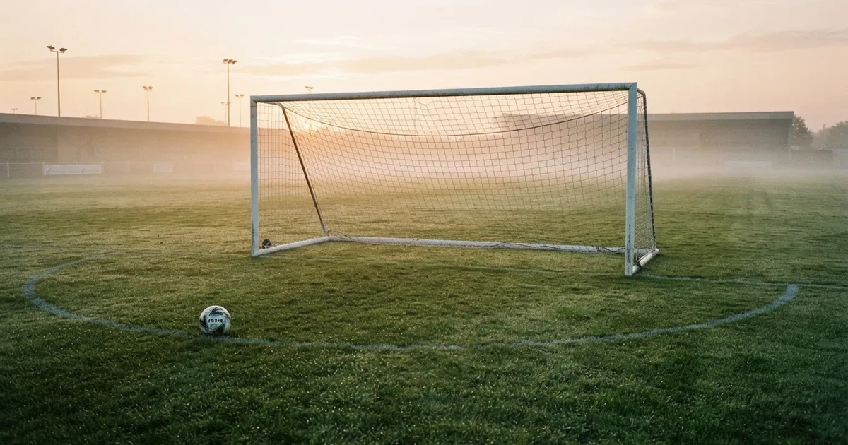 Ein Fußballtor mit gespanntem Netz steht auf einem leeren Rasenplatz, davor ein einzelner Ball, Morgendunst liegt über dem Feld.