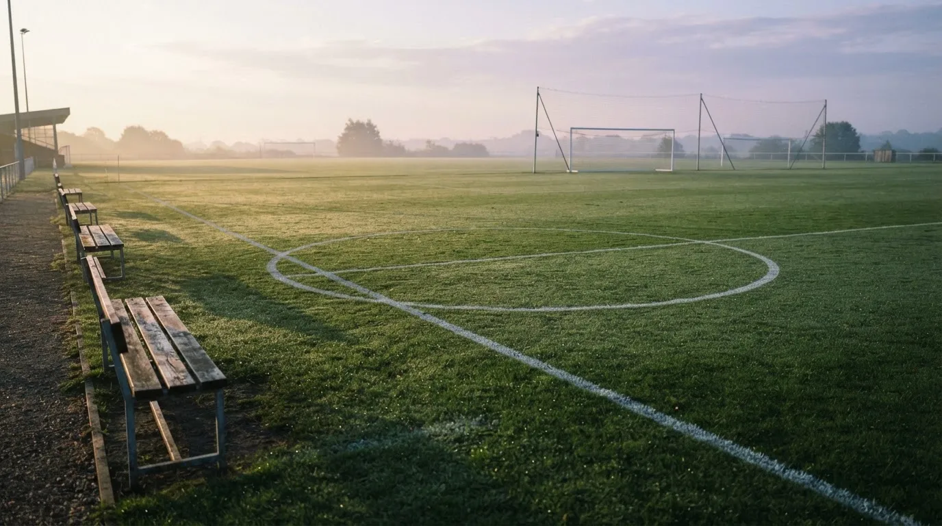 Ein Fußballplatz mit frisch gezogenen Kreidelinien im ersten Morgenlicht, leere Ersatzbänke am Spielfeldrand.