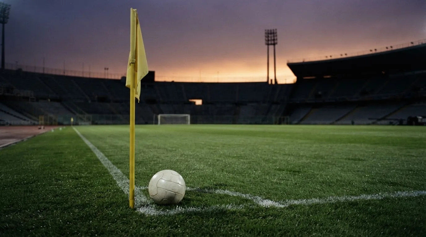 Ein Fußball liegt neben einem Eckfahnen-Pfosten auf einem Rasenplatz, im Hintergrund ein leeres Stadion in der Dämmerung.