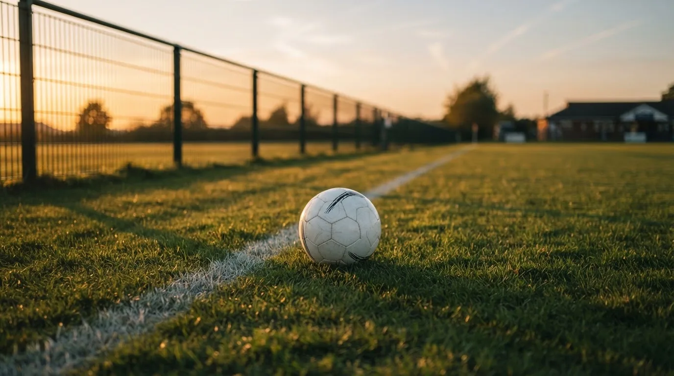 Ein Fußball liegt am Rand eines Rasens neben einer weißen Linie, im Hintergrund ein Zaun in der Abendsonne.