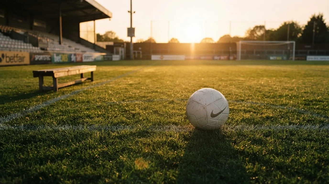 Ein Fußball auf einem Rasenplatz im Gegenlicht der tiefstehenden Sonne, am Spielfeldrand eine leere Ersatzbank.
