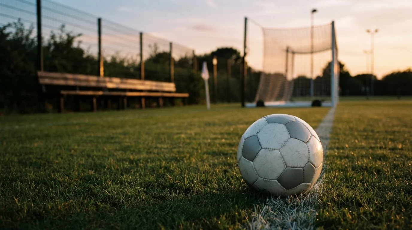 Ein Fußball liegt im Gras am Spielfeldrand, dahinter eine leere Bank und ein Tor im Gegenlicht.