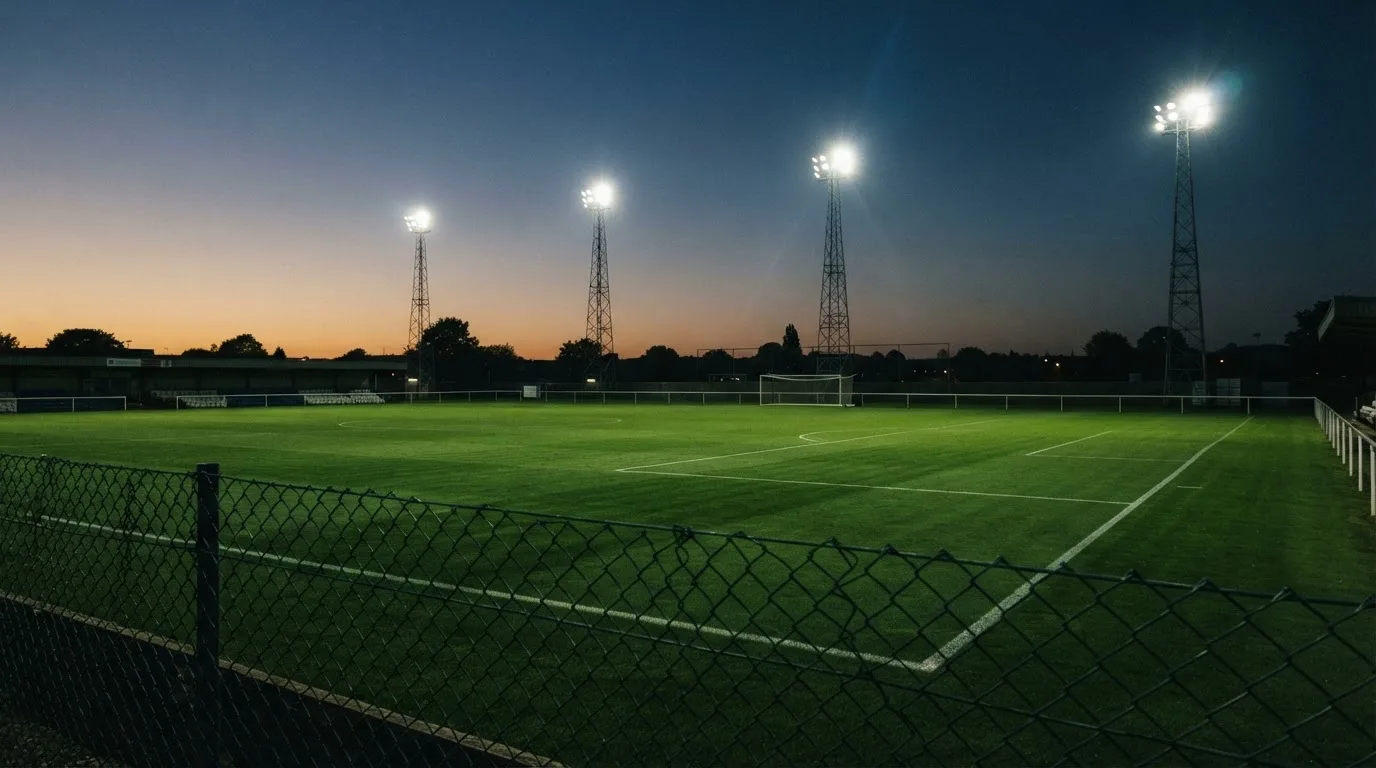 Eine Flutlichtanlage über einem leeren Sportplatz am Abend, im Vordergrund ein Maschendrahtzaun.