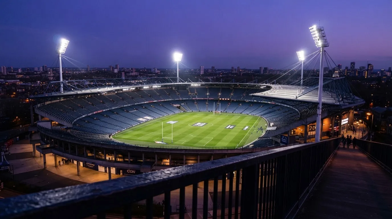 Ein Stadion bei Dämmerung mit eingeschalteten Flutlichtmasten, von einer Brücke aus aufgenommen.