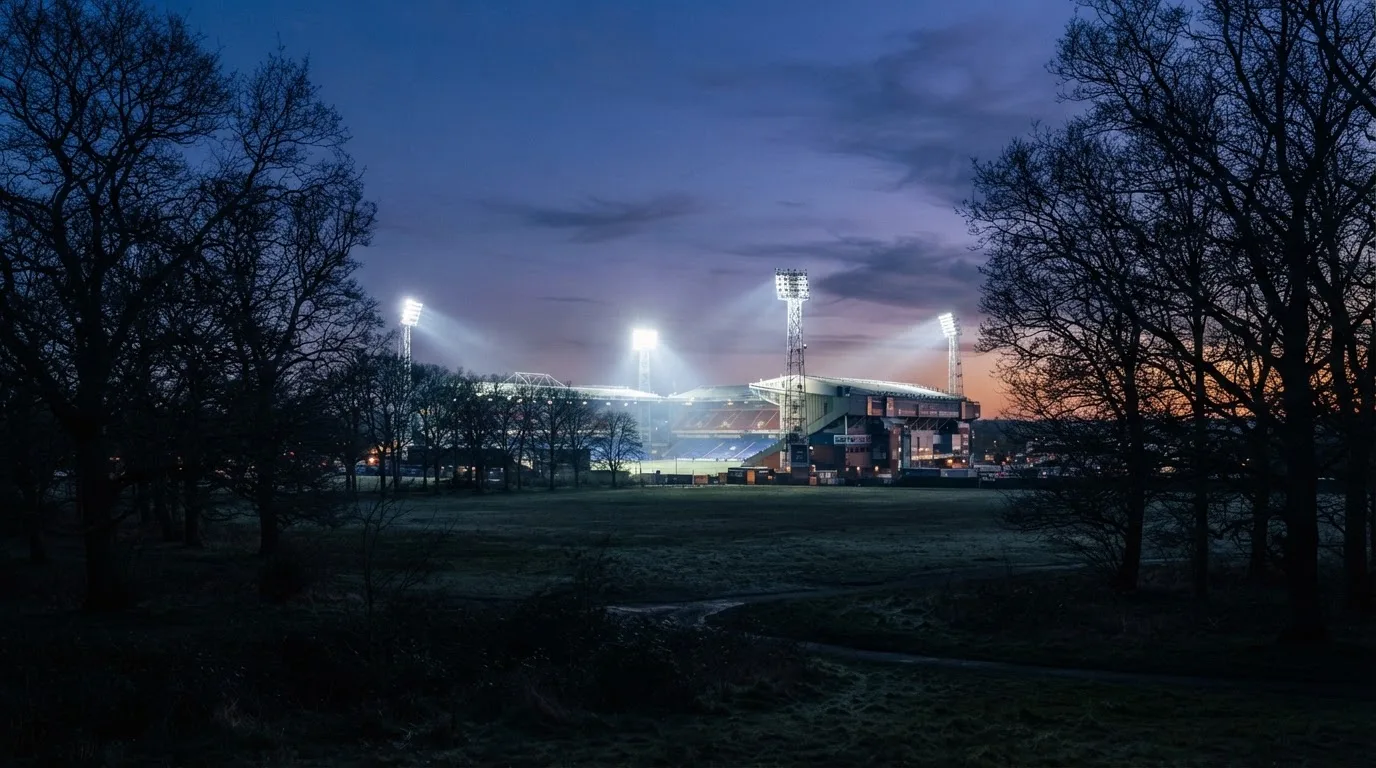 Ein Stadion unter Flutlicht aus großer Entfernung, davor die Silhouetten von Bäumen im Abendhimmel.