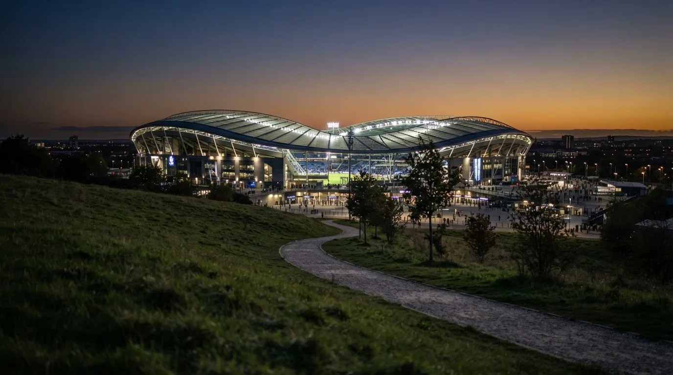Ein Stadion unter Flutlicht, von einer Anhöhe aus gesehen, davor ein stiller Weg in der Dämmerung.