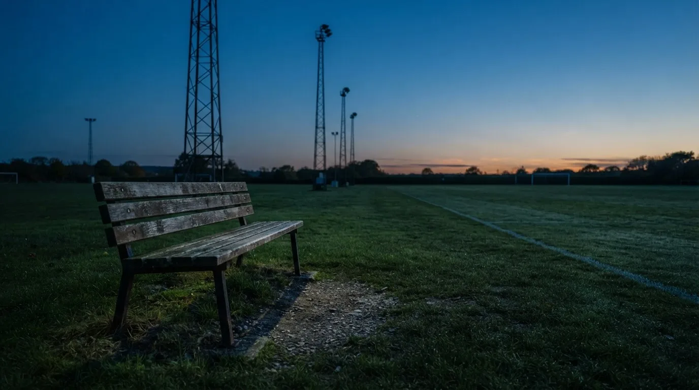 Eine leere Parkbank am Rand eines Fußballfelds in der Abenddämmerung, im Hintergrund ragen dunkle Flutlichtmasten in den Himmel.