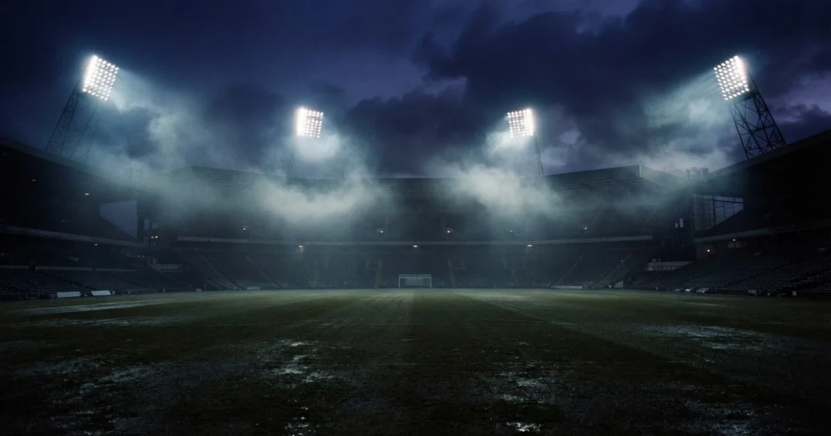 Ein Stadion unter Flutlicht, aus großer Entfernung über eine dunkle Wiese fotografiert, am Himmel zieht Nebel auf.