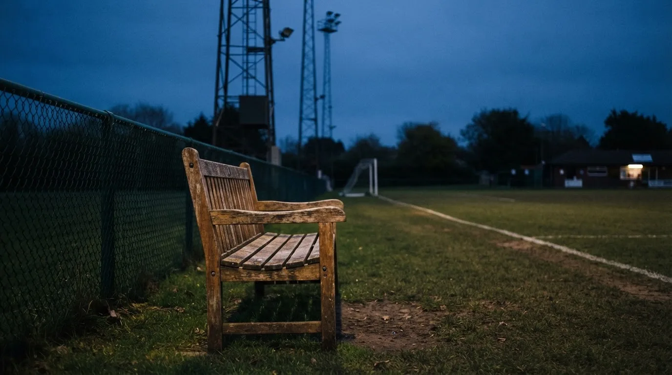 Eine leere Parkbank am Rand eines Fußballfelds in der Abenddämmerung, im Hintergrund ragen dunkle Flutlichtmasten in den Himmel.
