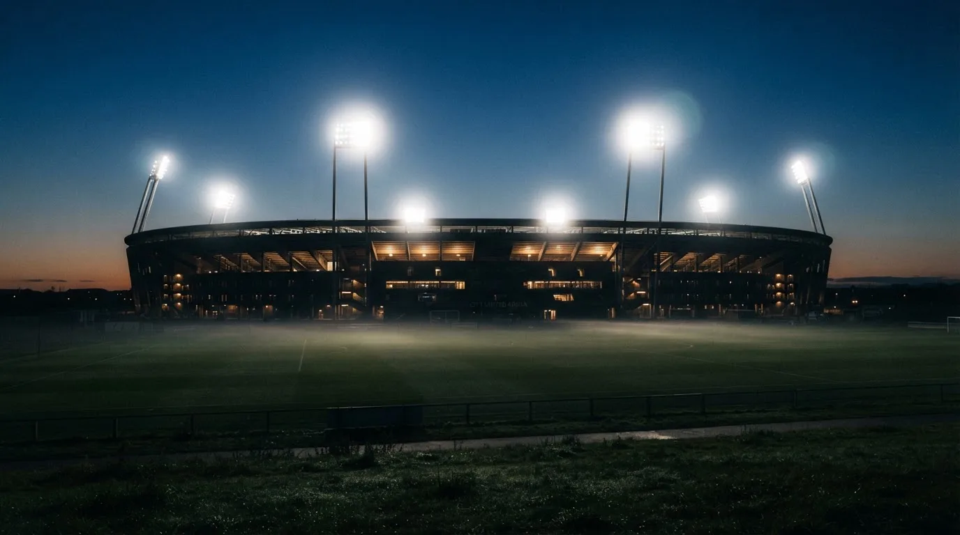 Ein großes Stadion unter Flutlicht, aus der Ferne über eine dunkle Wiese gesehen, die Lichtmasten spiegeln sich im Dunst der Abenddämmerung.