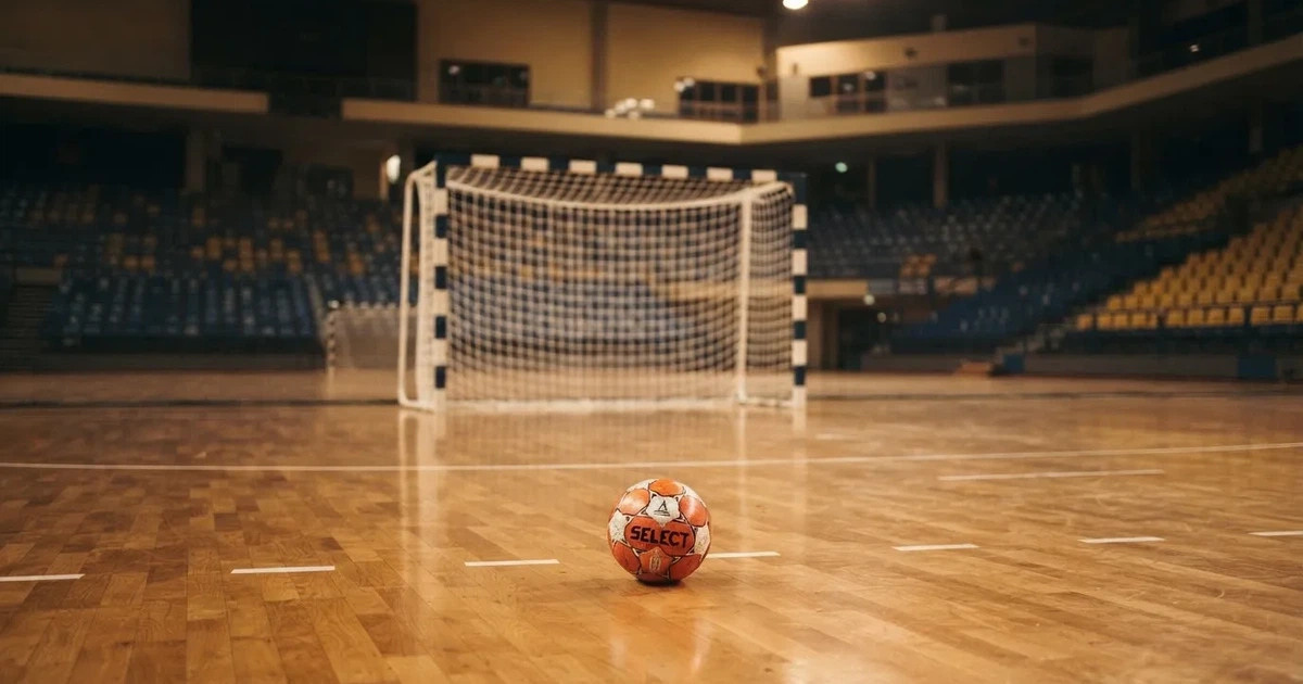 Ein Handball liegt auf dem Hallenboden vor einem Tor mit Netz, die leere Halle wird von Flutlicht beleuchtet.