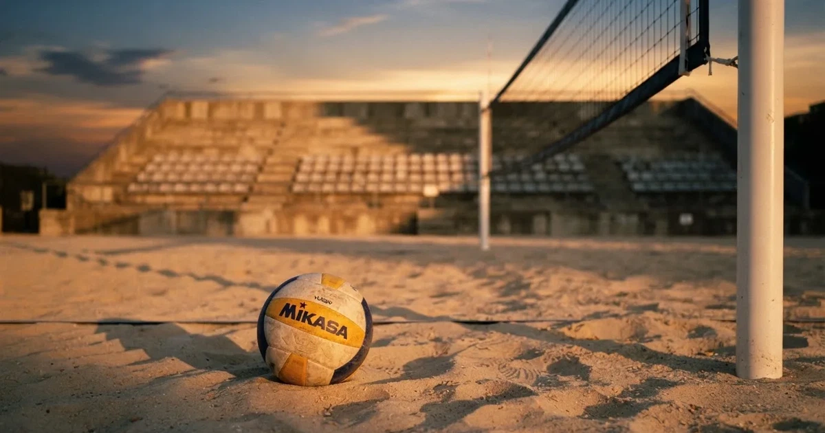 Ein Volleyball liegt auf dem Sand eines Beachvolleyball-Feldes neben dem gespannten Netz, im Hintergrund eine leere Tribüne bei Abendlicht.