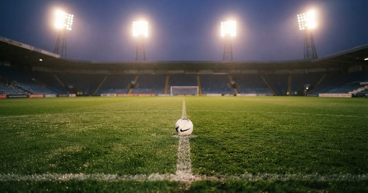 Ein Rasenplatz in der Dämmerung mit eingeschalteten Flutlichtmasten, ein Ball liegt am Anstoßpunkt, die Tribünen im Hintergrund sind unscharf.
