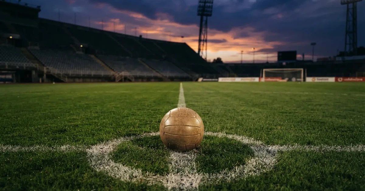 Ein Fußball liegt auf dem Anstoßpunkt eines leeren Rasenplatzes, im Hintergrund eine Tribüne und Flutlichtmasten in der Abenddämmerung.