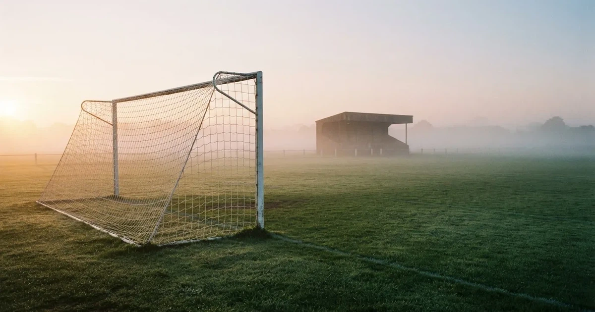 Ein Fußballtor mit Netz steht im Morgennebel auf einem leeren Rasenplatz, im Hintergrund die Silhouette einer kleinen Tribüne.