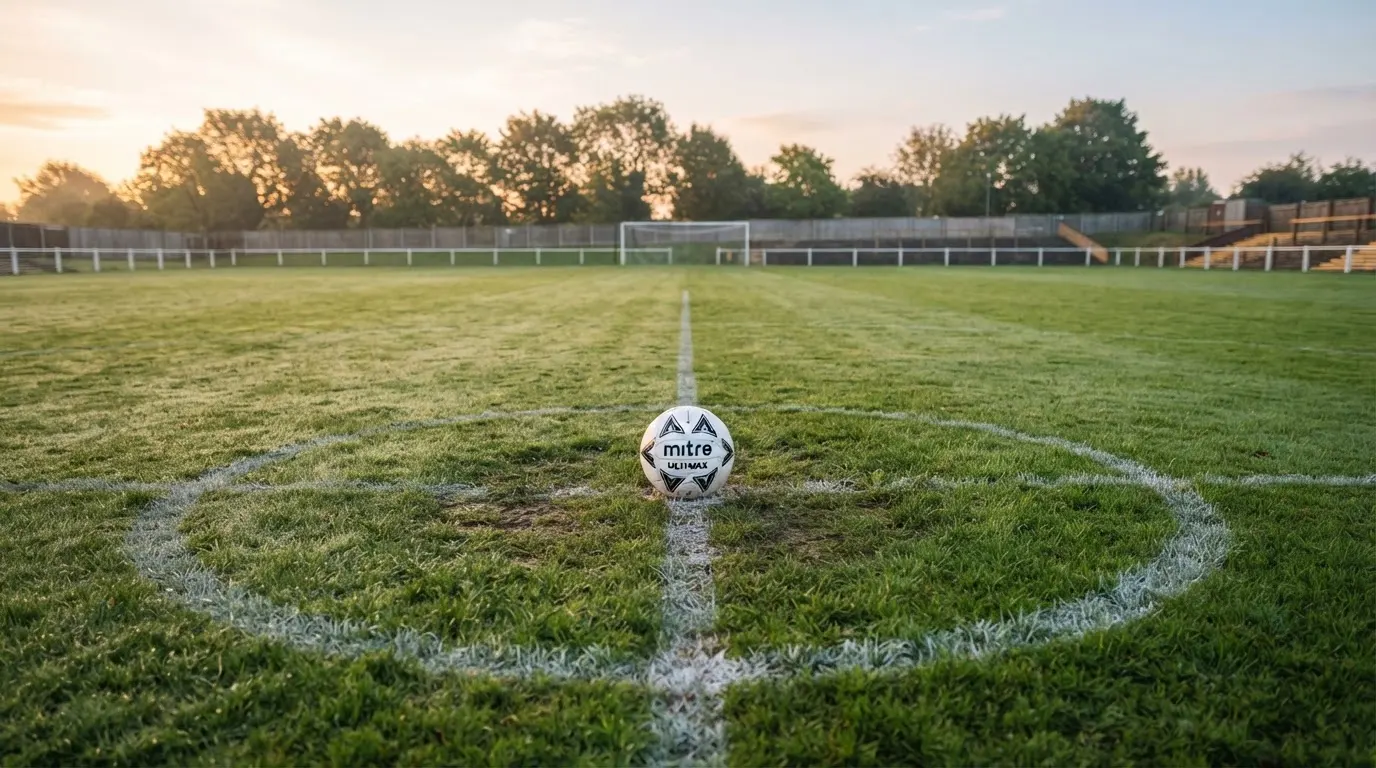 Ein Fußball liegt allein auf dem Anstoßpunkt eines leeren Rasenplatzes im Morgenlicht, am Rand ein niedriger Zaun.