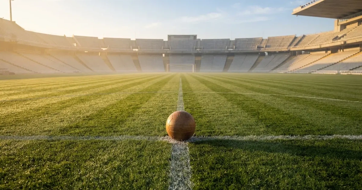 Ein Fußball liegt auf dem Anstoßpunkt eines Rasenplatzes, dahinter leere Tribünen im Licht des frühen Morgens.