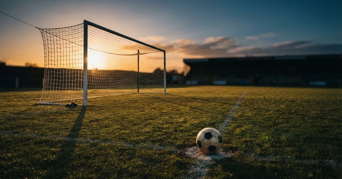 Ein Fußballtor mit Netz im Gegenlicht auf einem leeren Platz am Abend, davor ein Ball auf dem Rasen.
