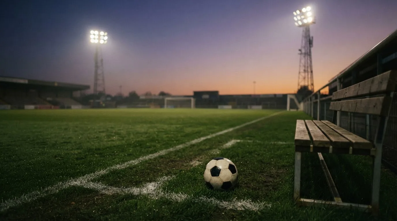 Ein Fußball liegt am Spielfeldrand neben einer leeren Ersatzbank, das Flutlicht über dem Platz leuchtet in der Dämmerung.