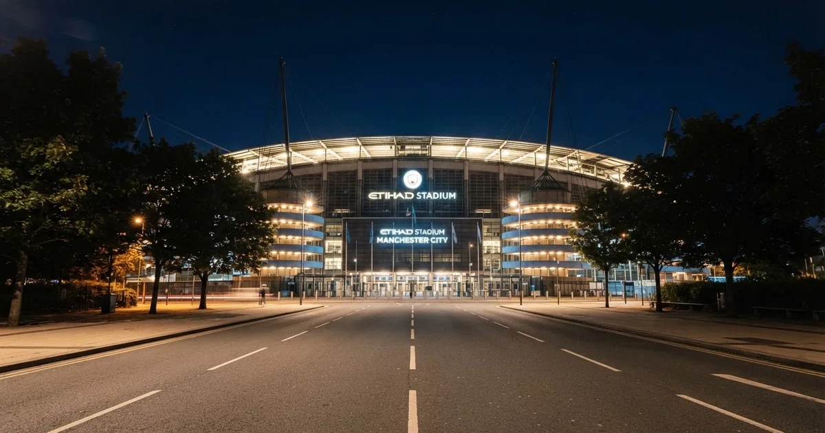 Ein hell erleuchtetes Fußballstadion bei Nacht aus der Ferne gesehen, über dem Dach ein klarer Himmel, davor eine leere Straße.
