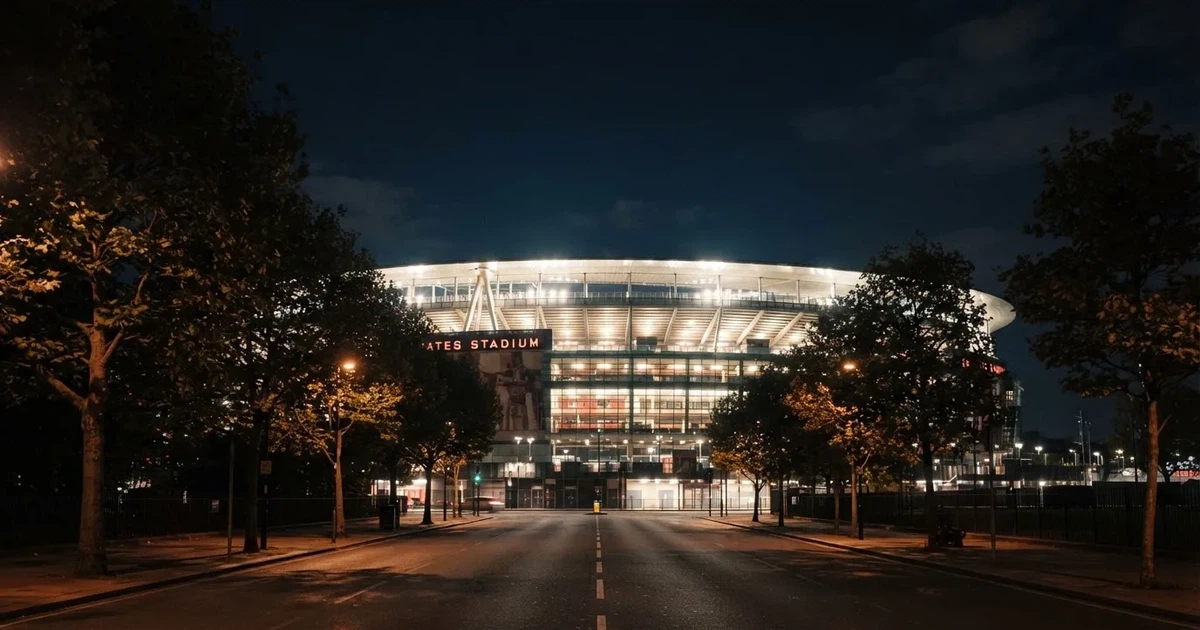 Ein hell erleuchtetes Stadion aus der Ferne bei Nacht, davor eine stille Straße und ein paar Bäume.