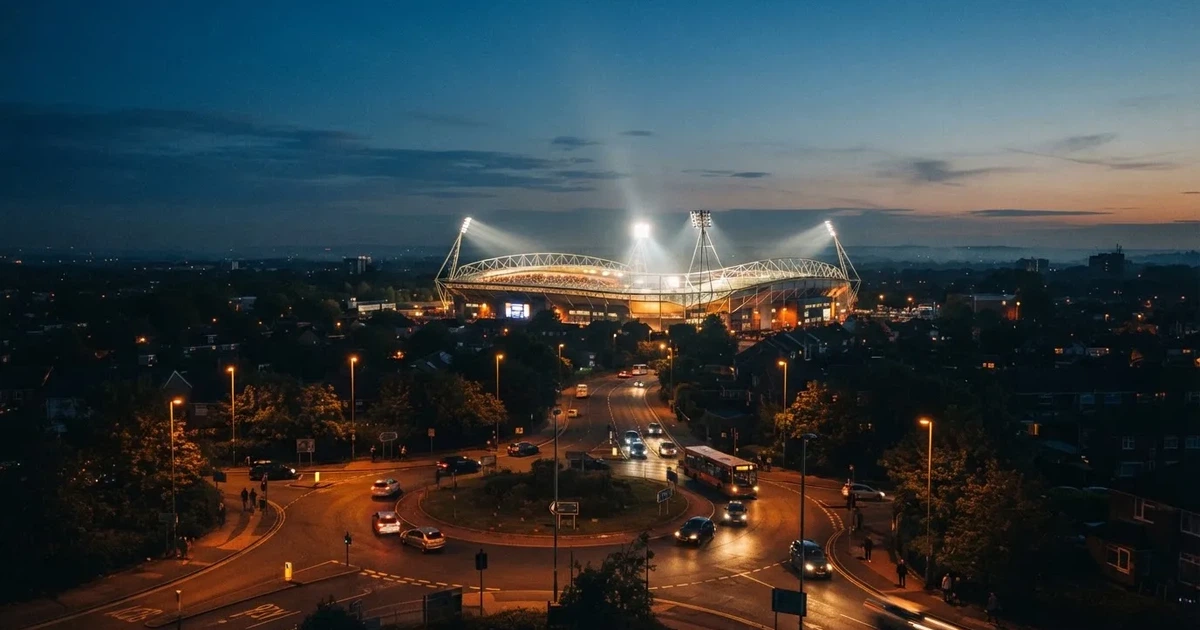 Ein Fußballstadion in der Dämmerung aus der Ferne, die Flutlichter gerade eingeschaltet, davor ein Kreisverkehr mit Laternen.
