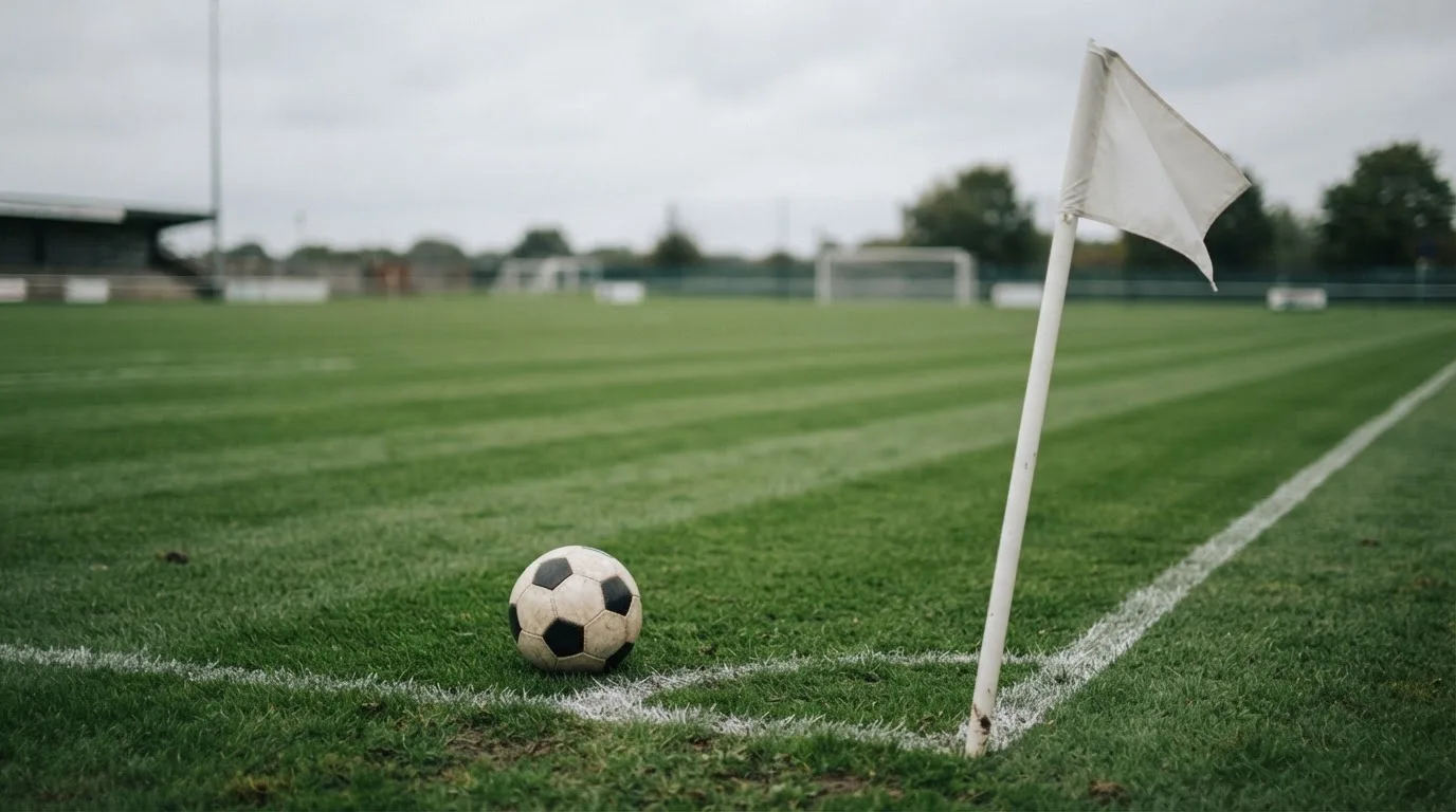 Ein Fußball liegt neben einer Eckfahne auf kurz gemähtem Rasen unter bewölktem Himmel.
